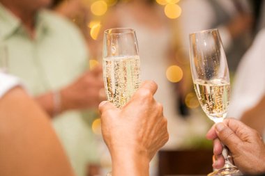 Düğünde şampanya bardaklarıyla tezahürat yapan bir grup insan. Gözlük ve ellere odaklanmış durumda. İnsanlar gülümsüyor ve mutlu görünüyorlar..