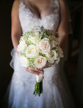 Beyaz gelinlik içinde güzel bir gelin elinde bir buket beyaz çiçek tutuyor. Gelin gülümsüyor, mutlu ve heyecanlı görünüyor. Buket güller, zambaklar ve şakayıklar dahil olmak üzere çeşitli beyaz çiçeklerden yapılmıştır. Gelin önümde duruyor.