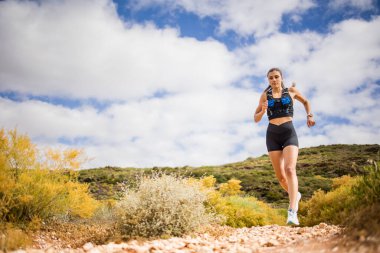 Female trail runner wearing black sportswear and a hydration vest, running through a scenic natural landscape on a rocky path. clipart