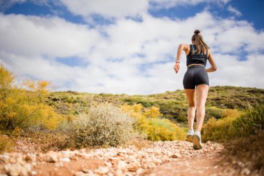Siyah spor giyim ve hidrasyon yeleği giyen kadın patika koşucusu kayalık bir yolda doğal bir manzarada koşuyor..