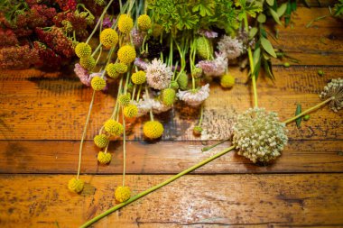 Rengarenk kır çiçekleri ve yaprakların kırsal bir düzeniyle ahşap bir masaya saçılmış doğal güzelliği ve çiçekli yaratıcılığı vurguluyor..