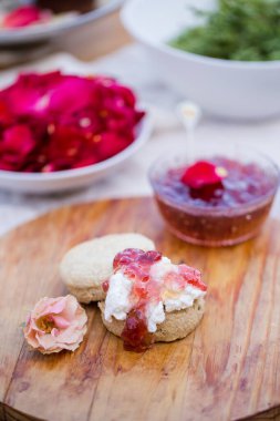 A freshly baked scone topped with clotted cream and rose petal jam, presented on a wooden board with a pink rose and bowls of rose petals and jam in the background. clipart