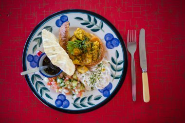 An inviting South African-inspired meal featuring curry, sambal, yogurt, papadum, and a flavorful chutney served on a vibrant, patterned plate against a bold red background. clipart