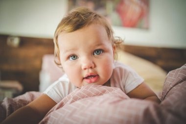 Mavi gözlü, sarı saçlı, yumuşak pembe yataklı, rahat, doğal ışıklandırılmış bir yatak odasında uzanan sevimli bir bebeğin yakın çekimi..