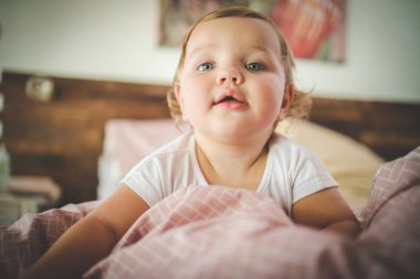 Mavi gözlü, sarı saçlı, yumuşak pembe yataklı, rahat, doğal ışıklandırılmış bir yatak odasında uzanan sevimli bir bebeğin yakın çekimi..