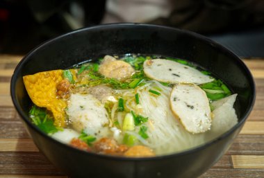 Siyah kasede servis edilen lezzetli Tayland erişte çorbasının yakın çekimi. Tabakta pirinç erişteleri, balık köfteleri, dilimlenmiş balık kekleri, gevrek wonton, taze otlar ve temiz aromalı et suyu yer alıyor. Orijinal Asya sokak yemekleri, geleneksel Tayland mutfağı konsepti.