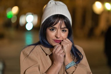 Smiling young female with makeup wearing white hat and brown coat standing on blurred night street and touching face while looking at camera