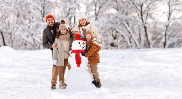 Happy parents and children gathering in snow-covered park together sculpting funny snowman from snow. Father, mother and two kids playing outdoor in winter forest. Family active holiday
