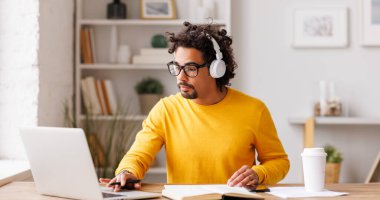 Gözlüklü ve kablosuz kulaklıklı siyahi bir adam masada otururken ve uzaktan çalışırken dizüstü bilgisayarda çalışıyor.