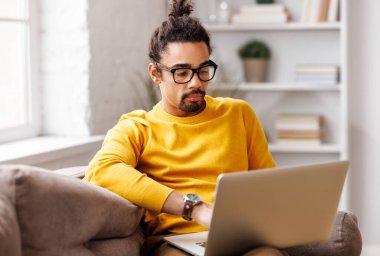 Gözlüklü Afrikalı Amerikalı bir adam, pencerenin yanındaki kanepede otururken, e-postaları ve sosyal ağları kontrol ediyor ve gündüzleri evden laptopta çalışıyor.