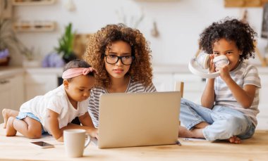 Genç sinirli Afro-Amerikan anne, evden iki küçük çocukla çalışırken kulaklarını kapatıyor ve laptop ekranına bakıyor, gürültücü çocuklar tarafından işten uzaklaştırılıyor.