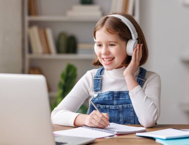 Günlük giysiler ve kablosuz kulaklıklar takan neşeli küçük bir öğrenci masada otururken ve evdeki çevrimiçi derste öğretmeniyle video görüşmesi yaparken gülümsüyor ve not defterine not alıyordu.