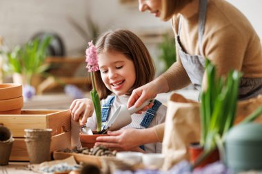 Önlüklü bir anne, mutlu kızıyla evde bahçeyle uğraşırken aynı zamanda dağınık bir masanın yanında durup hafta sonları birlikte bahçe işleriyle uğraşıyor.