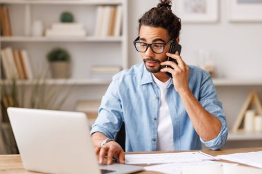 Genç, meşgul bir girişimci, evden bilgisayarla uzaktan arama yapıyor. Etnik adam uzaktan çalışma sırasında cep telefonuyla konuşuyor.