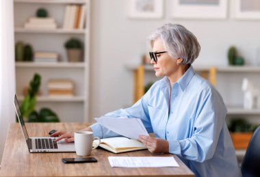 Bilgisayarda çalışan, belgeleri okuyan, notlar alan ve iç işlerinde çalışırken raporları analiz eden orta yaşlı kadın girişimci.