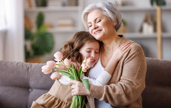 花の花束を持つ幸せなシニア祖母は優しく彼のかわいい孫娘を抱擁し お祝いと祖父母の日に休日を取得しながら彼女からの贈り物を受け取ります — ストック写真