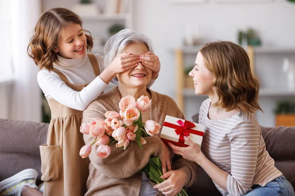 Happy International Women\'s Day. Smiling multi generational family daughter and granddaughter giving flowers  and gift to grandmother  cheerfully celebrate the spring holiday Mother\'s Day or grandparents day at home