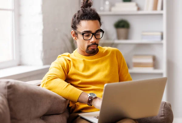African American man in glasses works, checks email and social networks  while sitting on sofa near window and working on laptop during work from home in daytime
