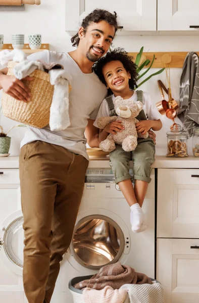 快乐的黑人爸爸 提着洗衣篮 微笑着 抱着乐观的孩子 看着相机 同时在家里的轻便厨房里做着家务活 — 图库照片