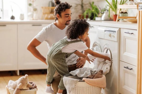 陽気な黒人の少年は家で週末のフライトキッチンで洗濯機の近くで洗濯しながらバスケットのリネンで父親を助けます — ストック写真