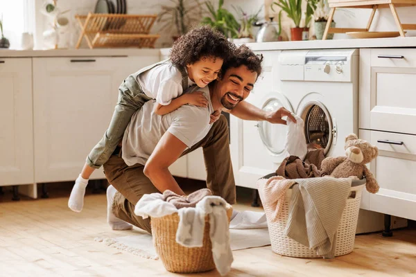 カーリーヘア笑顔のカジュアルな服で黒い子供は 家庭での朝のルーチン中にパパのローディング洗濯機を受け入れる — ストック写真