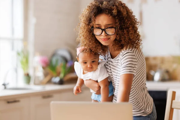 Multitasking young african american woman, working mother freelancer  using laptop computer with baby girl in hands, ethnic female doing freelance work on maternity leave