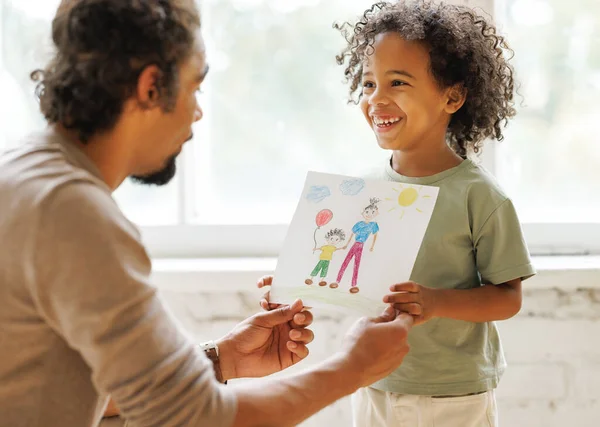 Happy Fathers Day. Cute little african american child son congratulating dad and giving him drawing with happy family on paper, smiling daddy and kid celebrating holiday at home