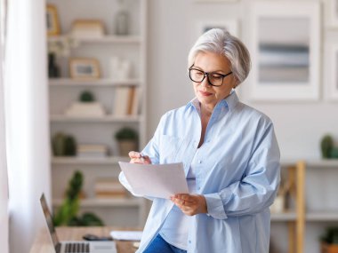Bilgisayarda çalışan, belgeleri okuyan, notlar alan ve iç işlerinde çalışırken raporları analiz eden orta yaşlı kadın girişimci.