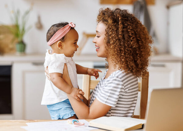 Freelance and motherhood. Happy young african american woman, self employed mother enjoying remote work from home with cute baby girl infant while sitting together in front of laptop 