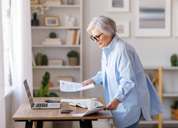 Focused middle aged female entrepreneur working at laptop, reading documents,  take notes and analyzing reports while working on project in domestic workplace