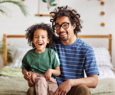 Neşeli Afro-Amerikan aile babası ve çocuk evde yatakta oturup kameraya bakıyor. Baba ve oğul..