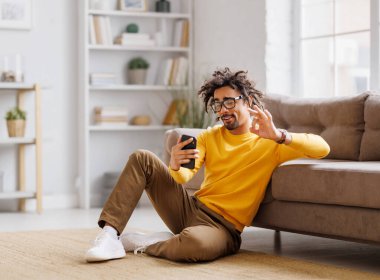 Sarı eşofmanlı ve gözlüklü mutlu Afrikalı Amerikalı adam. Evde, kanepede dinlenirken cep telefonunda gülümseyen ve sosyal medyada gezinen.