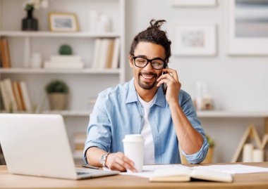 İçerik Afrikalı Amerikalı erkek girişimci cep telefonu görüşmeleri sırasında projeyi tartışırken yerli ofiste paket kahveyle masada oturuyor