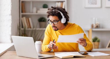 Gözlüklü, kablosuz kulaklıklı, laptopta kağıt okuma verisi olan siyahi bir adam masada otururken ve evden uzaktan çalışıyor.