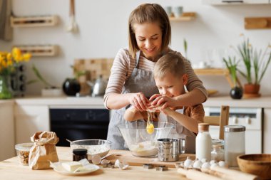 Content loving mother and cute boy adding ingredients in bowl and preparing dough at table at home clipart