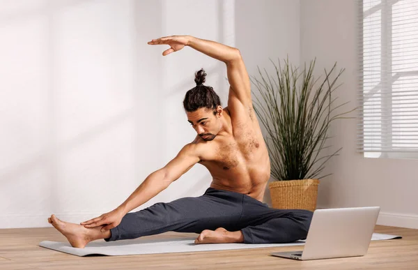 Shirtless African American man with hair bun raising arm and bending aside while sitting on mat near laptop and doing yoga in morning at home