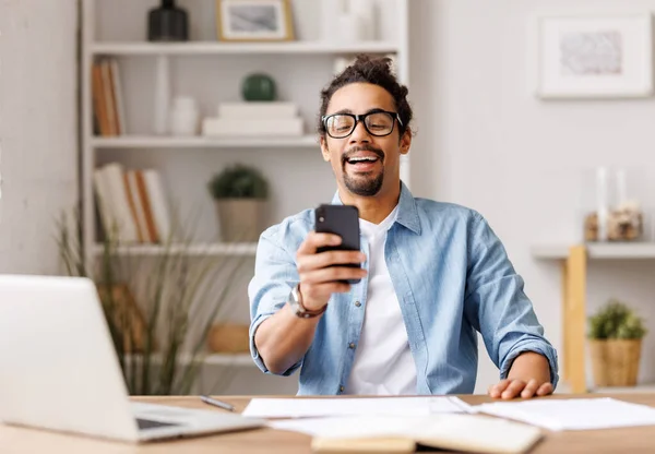 stock image Young african american male freelancer working on laptop computer at cozy home office, and browsing smartphone. Portrait of young ethnic man studying online. Freelance and distance education 