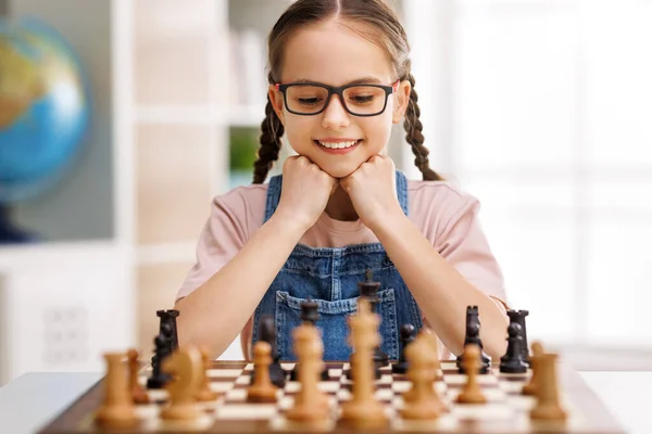 カジュアルな服で陽気な小さなプレーヤーが笑顔で 昼間のライト教室での試合中にチェスボードを見ています — ストック写真