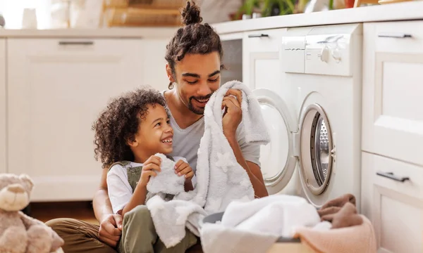 Neşeli zenci çocuk hafta sonu evde çamaşır makinesinin yanında çamaşır yıkarken çamaşır sepetinde babasına yardım ediyor.  