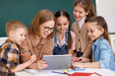 Mutlu bayan öğretmen ve iyimser çocuklar okulda ders sırasında masanın etrafında toplanırken tablette gülümseyip video izliyorlar.