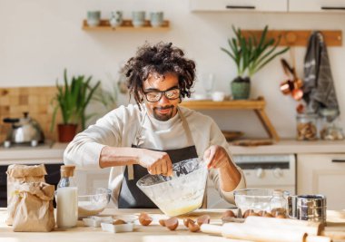 Önlüklü gülümseyen Afro-Amerikan erkek kaseye malzeme eklerken evde fırın için hamur yapıyor.