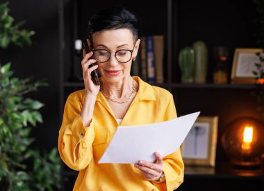Şık sarı bluz ve kağıt üzerinde veri okuyan gözlüklü olgun bir bayan yönetici ve iş sırasında cep telefonuyla müşteriyle konuşuyor.