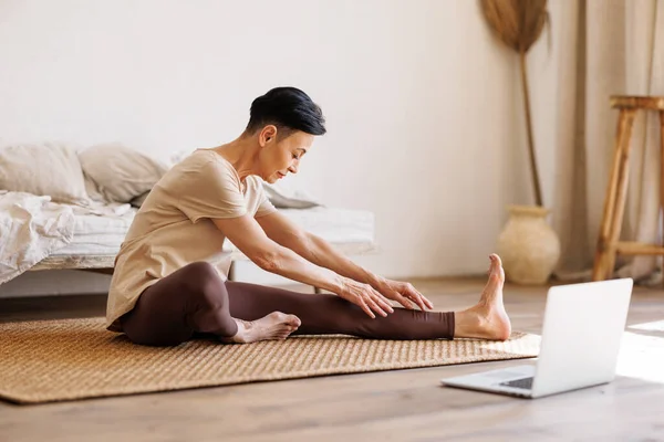 早上在家里进行在线训练时 穿着活动服的赤脚中年女性坐在床上和笔记本电脑之间 弯腰到腿上 — 图库照片