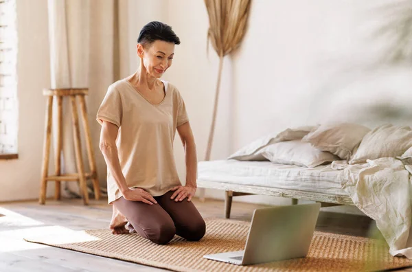 正成熟的女性 短短的黑发跪在地板上 靠近未铺好的床 早上在家里开始网上瑜伽课的时候看着笔记本电脑屏幕 — 图库照片