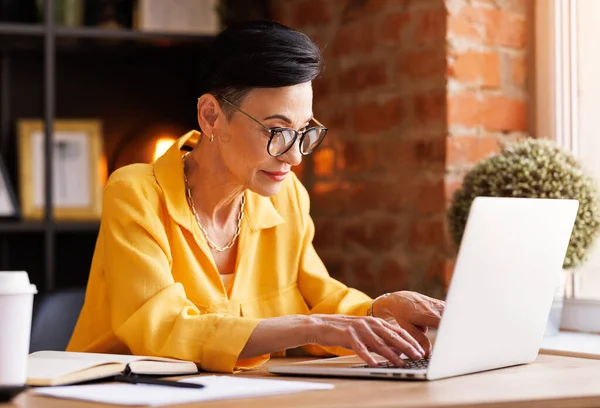 Middle aged female manager in stylish blouse with glasses typing on netbook keyboard while sitting at table and working on business project in home office with brick walls