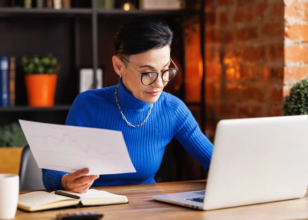 Mature businesswoman in blue turtleneck with glasses holding paper with graphs and looking at laptop while sitting at desk with laptop and working on business project in home office