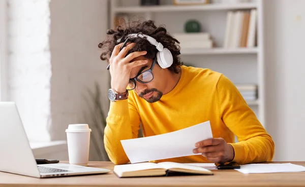 Frustrated tired young african american entrepreneur in glasses and headphones  suffering from headache with painful expression on her face, sitting in front of laptop  working remotely online. Frustrated black guy has problem