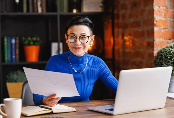 Mature businesswoman in blue turtleneck with glasses holding paper with graphs and looking at camera while sitting at desk with laptop and working on business project in home office