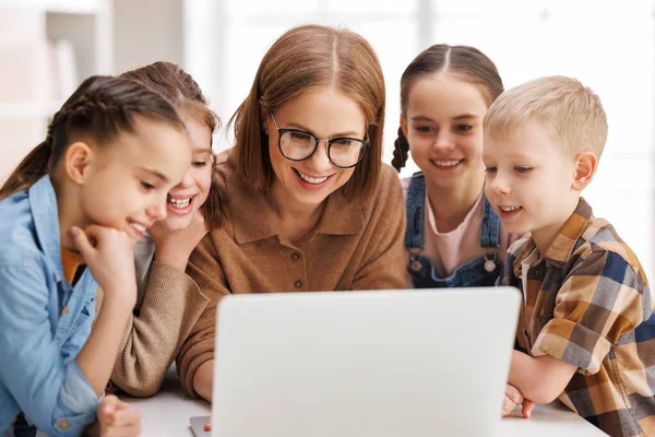 stock image Happy female tutor and group of schoolchildren smiling and watching educational video on laptop during lesson in sunlit classroom in school