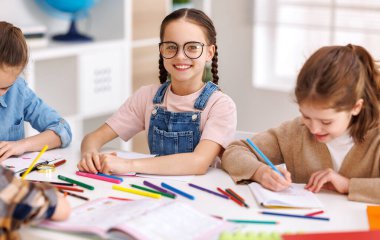 Gülümseyen ve kameraya bakan atkuyruklu neşeli okul çocuğu sınıf arkadaşlarının yanında oturuyor ve ders sırasında not defterine yazıyor.